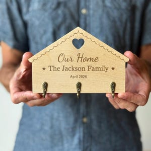 Könnte beinhalten: Ein hellfarbener, hausförmiger Schlüsselhalter aus Holz zur Wandmontage mit einem herzförmigen Ausschnitt an der Oberseite. Er trägt die Gravur Our Home, The Jackson Family und April 2026 sowie drei kleine bronzefarbene Metallhaken an der Unterseite.