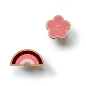May include: Two wooden drawer knobs with a natural light wood finish. One knob is shaped like a five-petaled flower with a pink center and a thin white outline. The other knob is shaped like a semi-circular rainbow with bands of dark red, pink, and light pink.