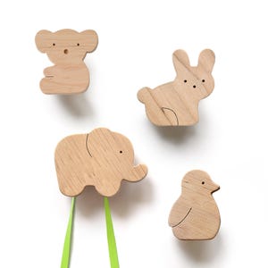 Puede incluir: Cuatro ganchos de pared de madera con forma de animales, cada uno con un diseño de animal diferente: un koala, un conejo, un elefante y un pingüino. El gancho con forma de elefante tiene dos cintas verdes.