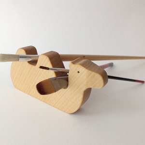 May include: A wooden camel-shaped brush holder with three paintbrushes in it. The holder is made of light wood and has a natural finish. The brushes are all different sizes and colors.