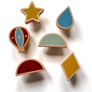 Könnte beinhalten: Ein Set aus sechs Holzknöpfen in verschiedenen Formen und Farben, darunter ein senfgelber Stern, ein blauer Regentropfen, ein rot-blauer Heißluftballon, ein salbeigrüner Halbkreis, ein dunkelroter Halbkreis und eine senfgelbe Rautenform.