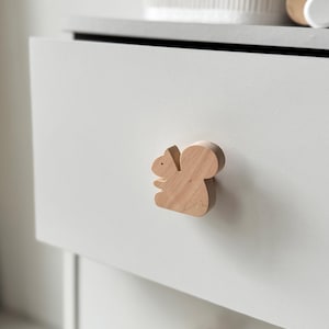 May include: A wooden drawer pull shaped like a squirrel. The squirrel is light brown and has a detailed design. The pull is attached to a white drawer.