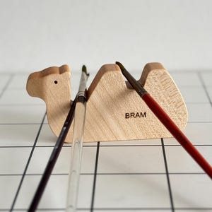 May include: Wooden camel brush holder with the word "BRAM" engraved on the side. Three paintbrushes are resting in the holder.