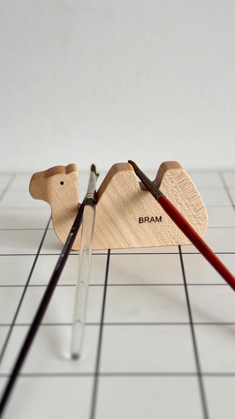 May include: Wooden camel brush holder with the word "BRAM" engraved on the side. Three paintbrushes are resting in the holder.