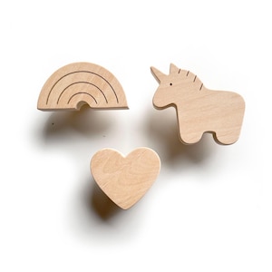 May include: Three wooden decorative shapes: a rainbow, a unicorn, and a heart. The rainbow has four curved lines. The unicorn has a horn and a small eye. The heart is a simple, rounded shape. All are made of light-colored wood.