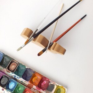 May include: A wooden paintbrush holder with four paintbrushes, including a black, white, and brown brush. A watercolor paint palette with 18 different colors is also shown.