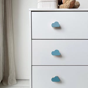 May include: White three-drawer chest of drawers with blue cloud-shaped handles. A stuffed bunny and a small, striped box sit on top. The chest is in a room with a sheer curtain.