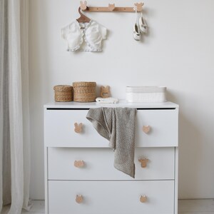 Puede incluir: C&oacute;moda blanca con tres cajones y pomos con forma de animales. La c&oacute;moda tiene una parte superior blanca con dos cestas de mimbre y una caja rectangular blanca. Un estante de madera con ganchos con forma de animales est&aacute; montado en la pared sobre la c&oacute;moda. Una chaqueta blanca y un par de zapatos blancos cuelgan de los ganchos.