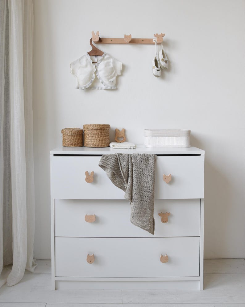 Puede incluir: C&oacute;moda blanca con tres cajones y pomos con forma de animales. La c&oacute;moda tiene una parte superior blanca con dos cestas de mimbre y una caja rectangular blanca. Un estante de madera con ganchos con forma de animales est&aacute; montado en la pared sobre la c&oacute;moda. Una chaqueta blanca y un par de zapatos blancos cuelgan de los ganchos.
