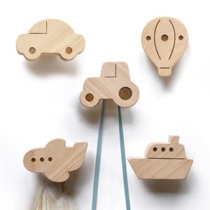 Peut inclure: Ensemble de cinq patères murales en bois en forme de voiture, tracteur, montgolfière, avion et bateau. Les crochets sont en bois clair et montés sur fond blanc. Parfaits pour une chambre d'enfant, ces crochets sont décoratifs et pratiques.