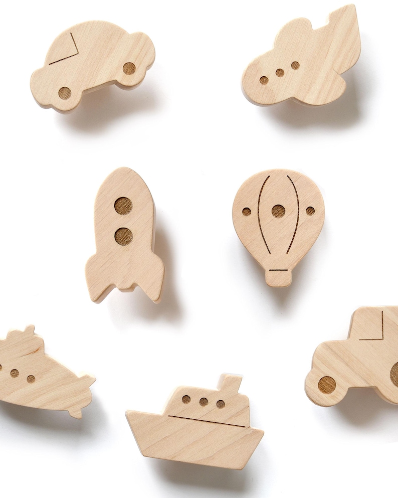 Individual wooden wall hooks for a boy's nursery featuring transport shapes: a rocket, car, plane, ship, submarine, and hot air balloon. These handcrafted vehicle-themed coat hooks are made from natural wood with engraved details. Displayed on a white background, sold per piece for custom wall decor.