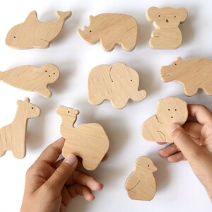Pode incluir: Uma coleção de figuras de animais em madeira de cor clara, incluindo uma baleia, rinoceronte, koala, foca, elefante, hipopótamo, girafa, camelo, leão e um pássaro. Cada peça apresenta um acabamento liso em madeira natural com detalhes gravados.