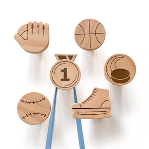 Puede incluir: Ganchos de pared de madera con forma de equipo deportivo, incluyendo un bate de béisbol, un balón de baloncesto, un disco de hockey, un patín de hielo, una mano con un guante y una medalla con el número "1" en ella.
