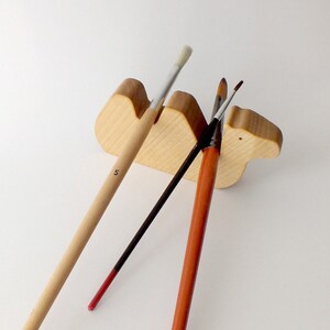 May include: Wooden camel brush holder with three paintbrushes. The brushes are a natural wood color, black, and red.