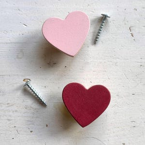May include: Two heart-shaped wooden decorative pieces, one light pink and one burgundy, are displayed on a white, textured surface. Two silver screws are also present. The hearts are likely for home decor or craft projects.