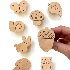 Puede incluir: Colección de formas de madera con temática de bosque, que incluye una mariposa, una seta, una hoja, un caracol, un conejo, una bellota, un erizo y un búho. Cada forma está tallada en madera clara y presenta detalles grabados.