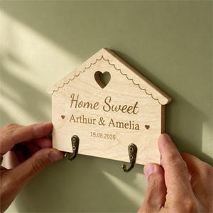 May include: A light-coloured wooden wall-mounted key holder shaped like a house with a scalloped roof edge and a heart cutout. It features the engraved text "Home Sweet", "Arthur & Amelia", and "15.08.2025" with two bronze-toned metal hooks.