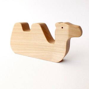 May include: A wooden camel toy, carved from light wood. The camel is lying down with its head turned to the side. The toy is simple and unpainted.