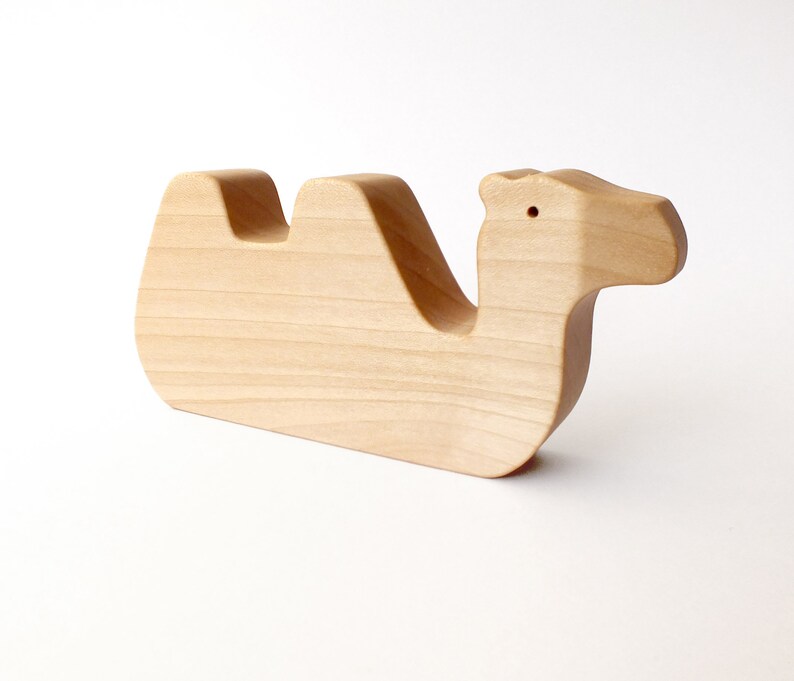 May include: A wooden camel toy, carved from light wood. The camel is lying down with its head turned to the side. The toy is simple and unpainted.