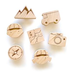 Könnte beinhalten: Eine Sammlung von Holzspielzeugen zum Thema Camping, darunter ein Berg, ein Wohnmobil, eine Karte, eine Kamera, ein Kompass und zwei Figuren mit Hüten. Die Spielzeuge sind aus hellem Holz mit gravierten Details gefertigt.