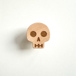 Pode incluir: Um item decorativo em forma de caveira de madeira. A caveira tem duas órbitas oculares circulares e uma boca retangular. O item é feito de madeira de cor clara com órbitas oculares castanhas mais escuras. A caveira está centrada em um fundo branco.