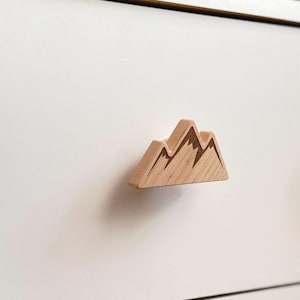May include: A wooden mountain-shaped drawer pull. The drawer pull is light brown with etched details depicting mountain peaks. It is mounted on a white drawer front, providing a natural accent to the furniture.