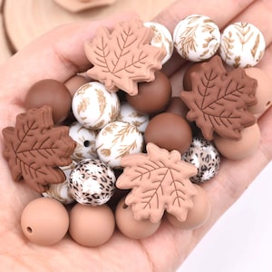 May include: A collection of silicone beads in various shapes and patterns, including solid matte spheres in shades of brown and tan, maple leaf shapes in light brown, and white spheres featuring gold botanical prints and brown leopard spot patterns.