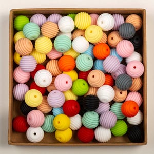 May include: A square cardboard box filled with a collection of round, ribbed silicone beads in a variety of colours including yellow, pink, orange, green, blue, white, black, and grey. Each bead features a central hole for threading purposes.