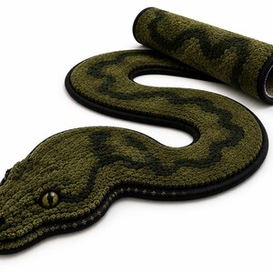 Puede incluir: Una alfombra con forma de serpiente con una superficie texturizada y copetuda en tonos verde oliva y verde oscuro. La alfombra presenta un borde negro, una cabeza detallada con un ojo amarillo prominente y un cuerpo largo y sinuoso.