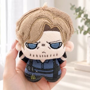 May include: A small plush toy featuring a character with light brown hair, blue eyes, and a serious expression. The figure wears a blue shirt with black tactical straps and a small green pouch detail on the waist, crafted from soft fabric materials.