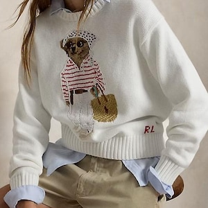 May include: A white long-sleeve knit sweater featuring a graphic of a bear wearing a polka dot headscarf, a red and white striped shirt, and white pants, while carrying a tan woven bag. The letters RL are embroidered in red on the lower left side.