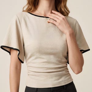 May include: A beige short-sleeved top featuring a boat neckline with black trim, flared sleeves with black edges, and gathered side detailing at the waist. The garment is paired with dark gray trousers and a gold necklace and bracelet.