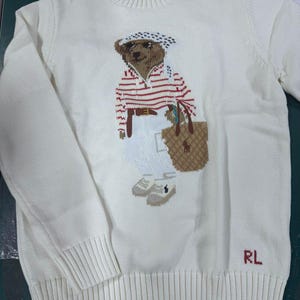 May include: A cream-colored crewneck sweater featuring a knit graphic of a bear wearing a red and white striped shirt, white pants, and a patterned headscarf, while carrying a tan tote bag. The letters RL are embroidered in red at the bottom.