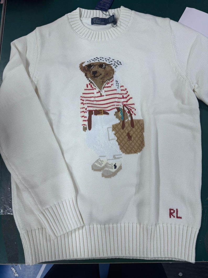 May include: A cream-colored crewneck sweater featuring a knit graphic of a bear wearing a red and white striped shirt, white pants, and a patterned headscarf, while carrying a tan tote bag. The letters RL are embroidered in red at the bottom.