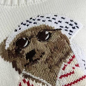 May include: A white knit sweater featuring an embroidered bear graphic. The bear is brown with a black nose, wearing round black sunglasses and a white patterned head covering. The bear is dressed in a white shirt with thin red horizontal stripes.