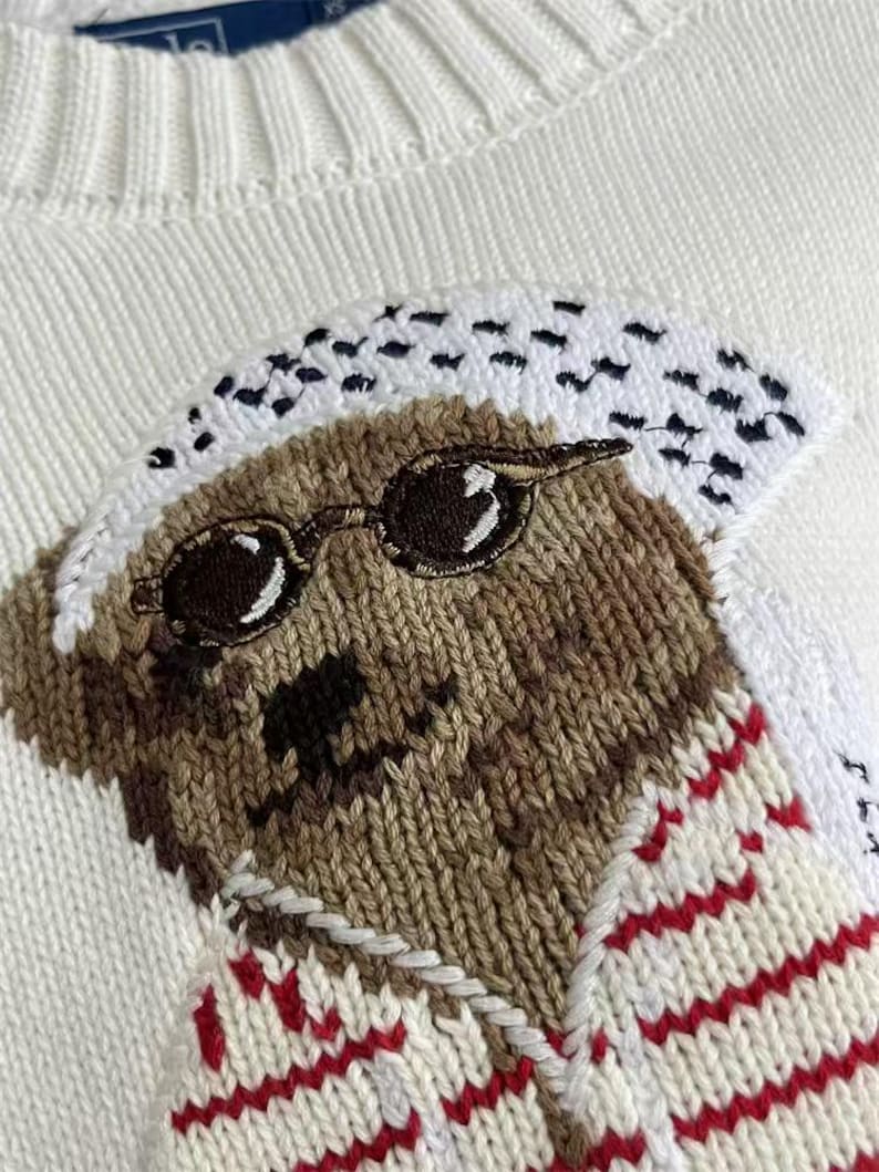May include: A white knit sweater featuring an embroidered bear graphic. The bear is brown with a black nose, wearing round black sunglasses and a white patterned head covering. The bear is dressed in a white shirt with thin red horizontal stripes.