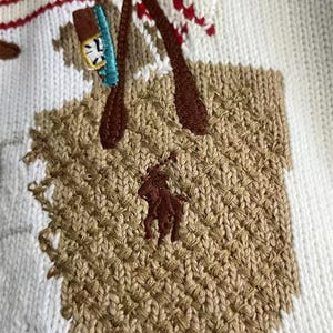 May include: A white knit sweater featuring an embroidered tan tote bag with dark brown handles. The bag displays a dark brown polo player logo and contains a small blue and white object. Red and white striped patterns appear above the bag.
