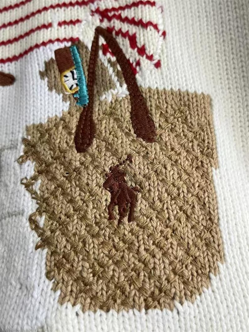 May include: A white knit sweater featuring an embroidered tan tote bag with dark brown handles. The bag displays a dark brown polo player logo and contains a small blue and white object. Red and white striped patterns appear above the bag.