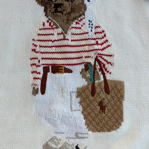 May include: A white knit sweater featuring an embroidered brown bear wearing a white and black patterned headscarf, sunglasses, a red and white striped long-sleeve shirt, a brown belt, white trousers, and light gray sneakers, carrying a tan tote bag.