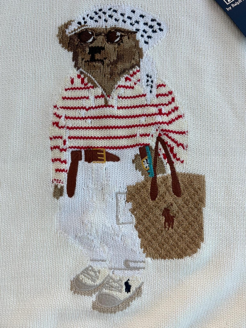 May include: A white knit sweater featuring an embroidered brown bear wearing a white and black patterned headscarf, sunglasses, a red and white striped long-sleeve shirt, a brown belt, white trousers, and light gray sneakers, carrying a tan tote bag.