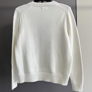 May include: A solid white long-sleeve sweater featuring a ribbed crew neckline, ribbed cuffs, and a ribbed hem. The garment is constructed from a knit fabric and is displayed on a black hanger, showcasing a clean, plain design with long sleeves.