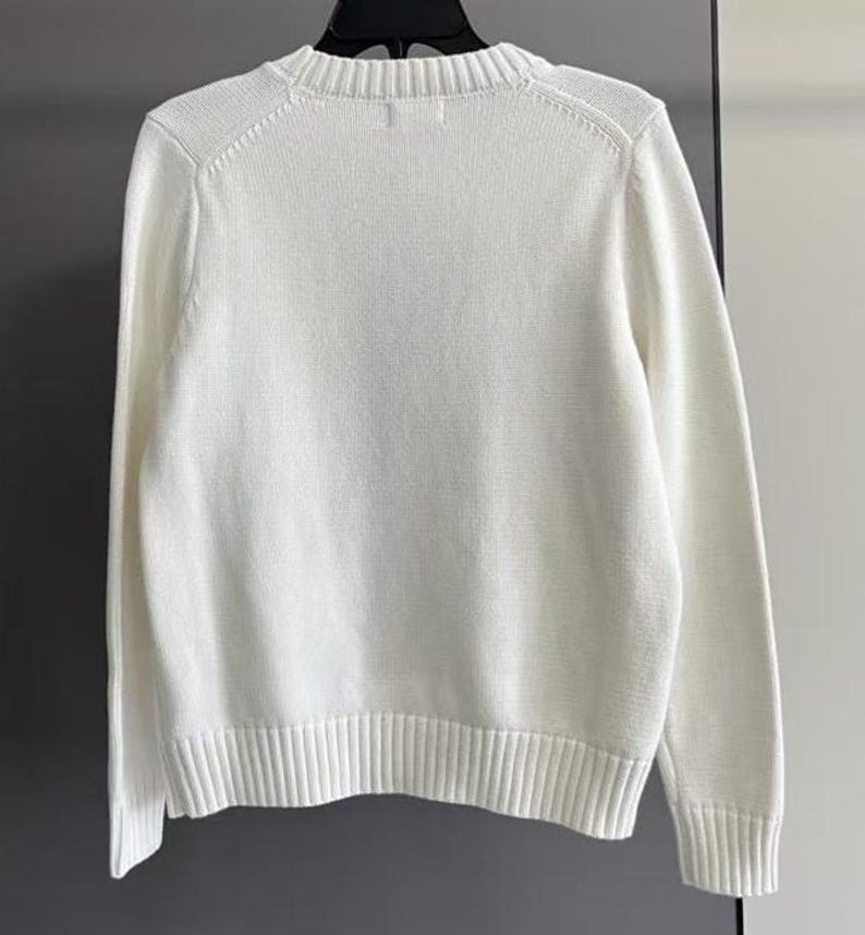 May include: A solid white long-sleeve sweater featuring a ribbed crew neckline, ribbed cuffs, and a ribbed hem. The garment is constructed from a knit fabric and is displayed on a black hanger, showcasing a clean, plain design with long sleeves.