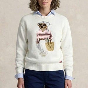 May include: A cream-colored long-sleeve knit sweater featuring a central graphic of a bear wearing a headscarf, sunglasses, and a striped shirt, carrying a woven tote bag. The letters RL are embroidered in red on the lower right hem.