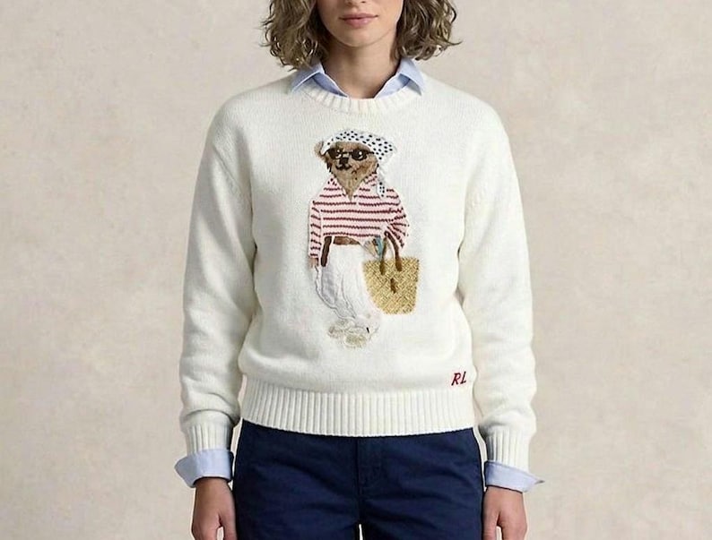 May include: A cream-colored long-sleeve knit sweater featuring a central graphic of a bear wearing a headscarf, sunglasses, and a striped shirt, carrying a woven tote bag. The letters RL are embroidered in red on the lower right hem.