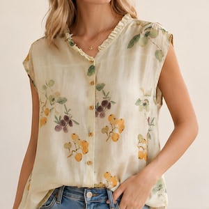 May include: A sleeveless button-down blouse in a light cream color featuring a delicate watercolor-style print of green leaves and clusters of yellow and purple fruit. The top has a small ruffled collar and light brown buttons down the front.