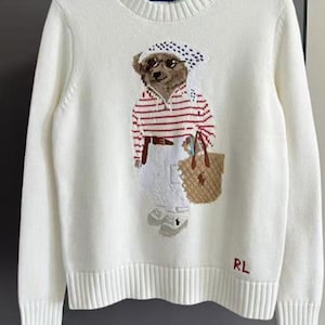 May include: A cream-colored long-sleeve knit sweater featuring a central graphic of a bear wearing a polka-dot headscarf, sunglasses, a red and white striped shirt, and white pants while carrying a tan tote bag. The letters RL are embroidered in red.