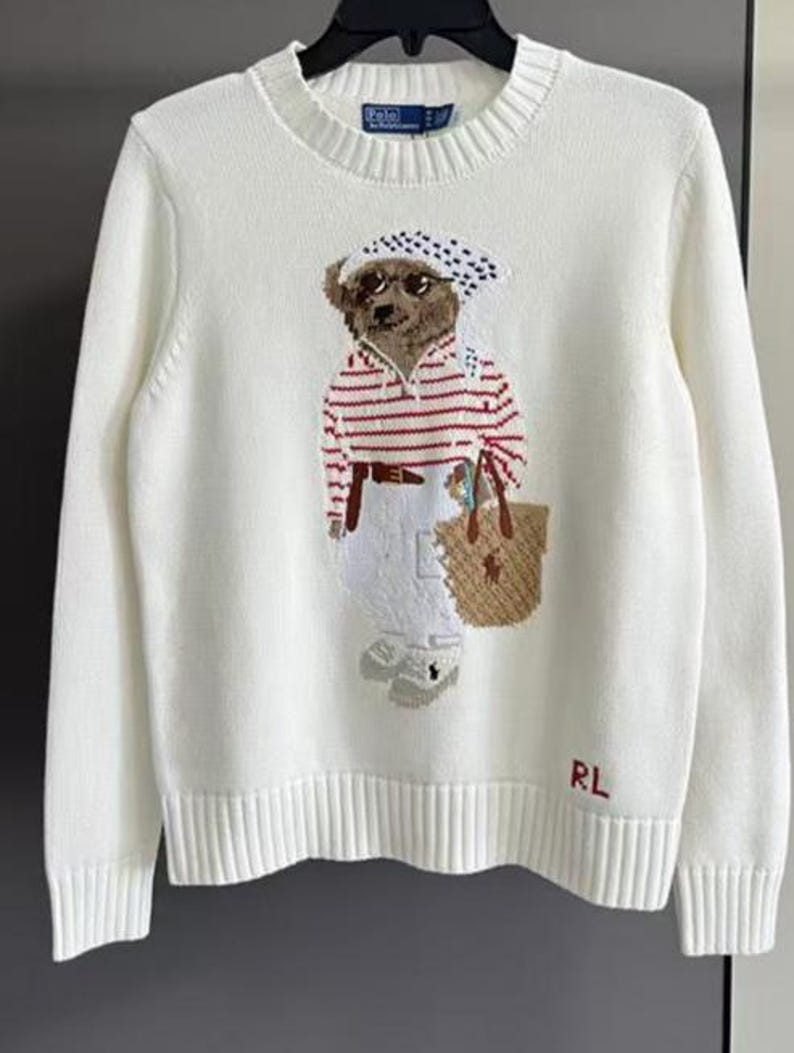 May include: A cream-colored long-sleeve knit sweater featuring a central graphic of a bear wearing a polka-dot headscarf, sunglasses, a red and white striped shirt, and white pants while carrying a tan tote bag. The letters RL are embroidered in red.