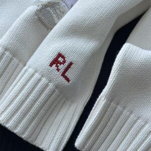 May include: A white knit sweater featuring ribbed cuffs and a red embroidered RL monogram on the sleeve. The fabric has a visible knit texture, and a portion of a white and tan patterned design is visible near the top of the sleeve.
