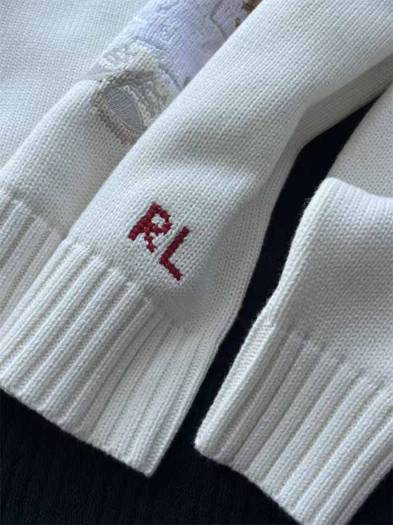 May include: A white knit sweater featuring ribbed cuffs and a red embroidered RL monogram on the sleeve. The fabric has a visible knit texture, and a portion of a white and tan patterned design is visible near the top of the sleeve.