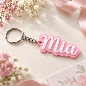 Op de afbeelding: Een sleutelhanger met een roze, rechthoekig label met geschulpte randen waarop de naam Mia in wit, verhoogd cursief schrift staat. Het label is met een kort kettinkje bevestigd aan een zilverkleurige metalen ring.